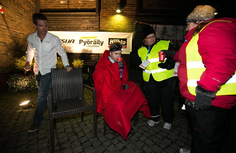 Vaarojen Maratonin vapaaehtoiset auttamassa osallistujaa illan h&auml;m&auml;r&auml;ss&auml; huoltopisteell&auml;, osallistuja k&auml;&auml;riytyneen&auml; l&auml;mpim&auml;&auml;n huopaan.