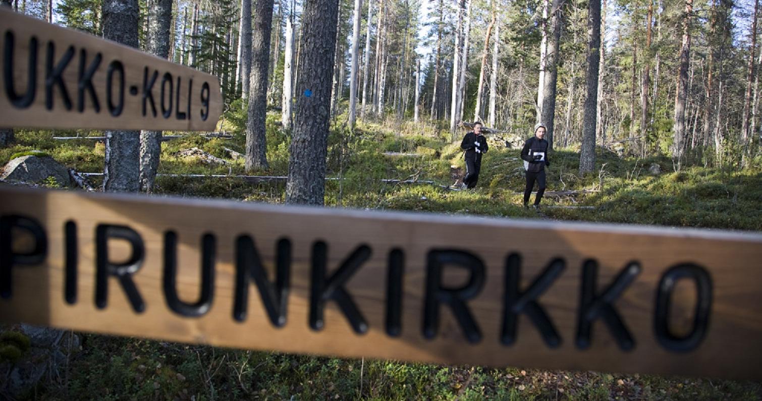 Vaarojen Maratonin reittimerkki 'Pirunkirkko', taustalla juoksijat etenevät Kolin kansallispuiston metsäpolulla kirkkaassa päivänvalossa.