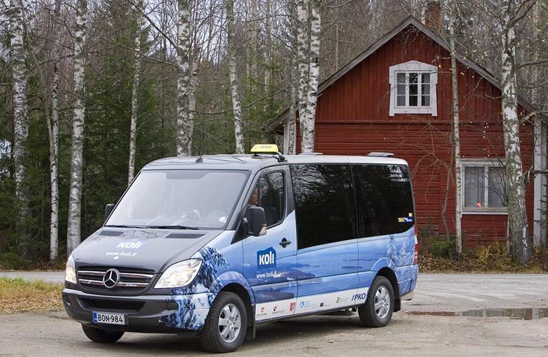 Sininen taksi-minibussi seisoo hiekkaparkissa punaisen m&ouml;kin edustalla, ymp&auml;rill&auml; lehtipuita syksyisess&auml; maisemassa.