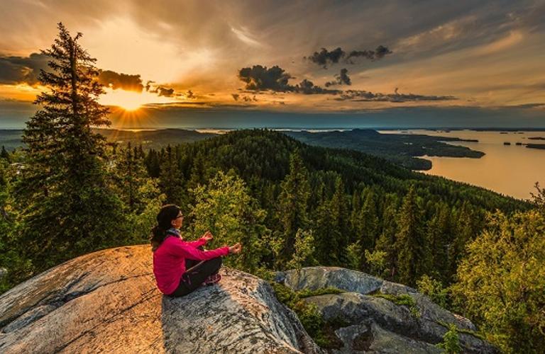 Nainen istuu Kolin huipulla katsellen laajoja j&auml;rvimaisemia ja v&auml;rikk&auml;it&auml; metsien latvoja ilta-auringon valossa.